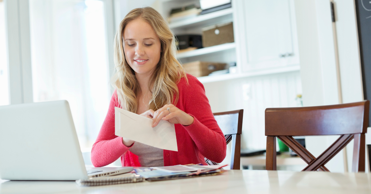 Young caucasian woman opening a donor acknowledgement letter while seated at a table with a laptop and stack of mail.