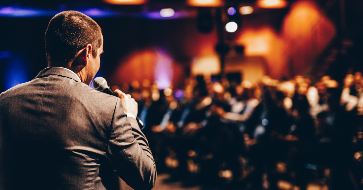 Image from behind a stage as a man speaks at an event.