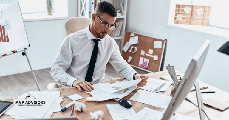 Man wearing a white shirt and black tie sits at a desk surrounded by papers and his computer, looking stressed. This image is the header for the blog about Time-Saving Fundraising Tips for Giving Season