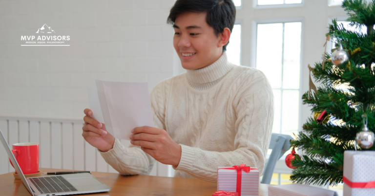A young man planning year-end fundraising campaigns looks at a giving season appeal letter while sitting at his computer and next to a holiday tree