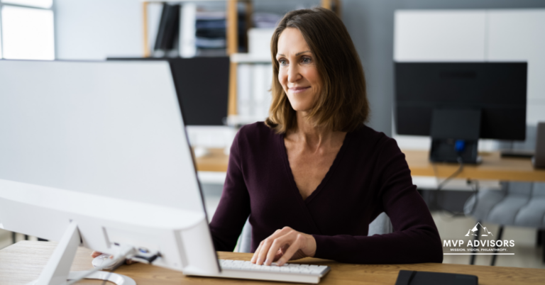 Year-End Fundraising Tools Blog header image - a woman sits at a desk in an office while working on a computer and smiling. Also includes the MVP logo