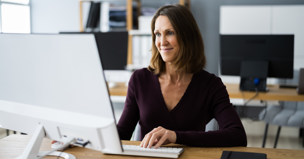 Year-End Fundraising Tools Blog header image - a woman sits at a desk in an office while working on a computer and smiling.